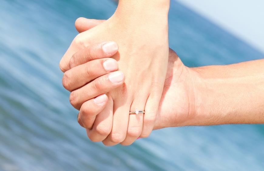 Manos de una pareja de novios comprometidos donde se ve el anillo de compromiso y al fondo el mar en la isla tabarca de Alicante
